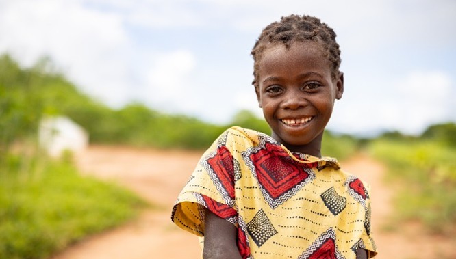 A girl in traditional clothing smiles against a rural background.